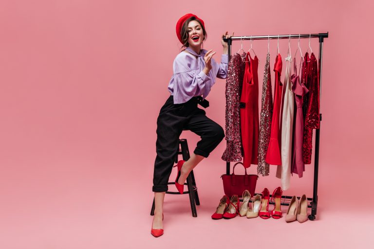 Slim lady in fashionable trousers, blouse and red hat is sitting on chair next to stand with shiny dresses