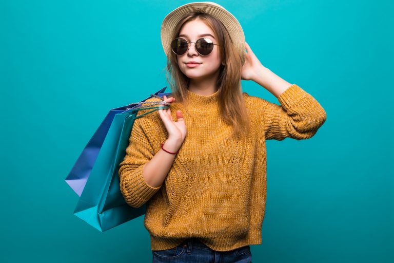 Young teen woman in sunglasses and hat holding shopping bags in her hands feeling so happiness isolated on green background